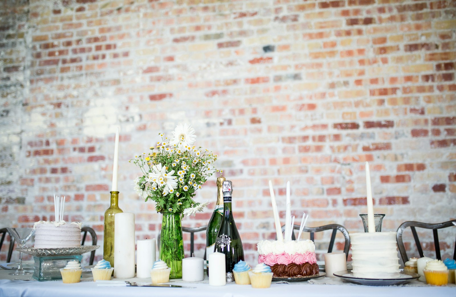 Styled celebration table with cakes, cupcakes, candles and fresh flowers against a brick wall